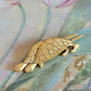 May include: A gold-toned brooch featuring two stylized turtles with detailed shell patterns. The turtles are facing the same direction and appear to be walking.