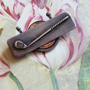 May include: A silver-toned golf club pin with a textured surface. The pin is shaped like a golf club with a ball at the end. The pin is on a floral background.