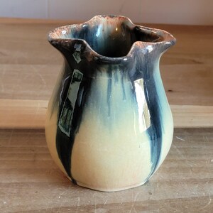 May include: A handmade ceramic vase with a unique, ruffled rim. The vase features a color gradient, transitioning from a creamy yellow base to a teal and dark blue top. The rim has a brown and orange accent. The vase is a decorative item.