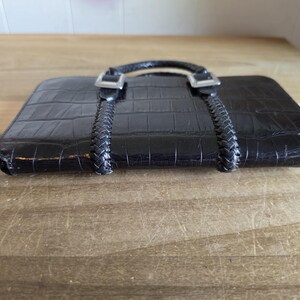 May include: A black rectangular wallet with a textured crocodile skin pattern. The wallet features two braided black straps extending from the top, secured by silver-toned metal buckles. A small, dark, heart-shaped zipper pull is attached to the side.