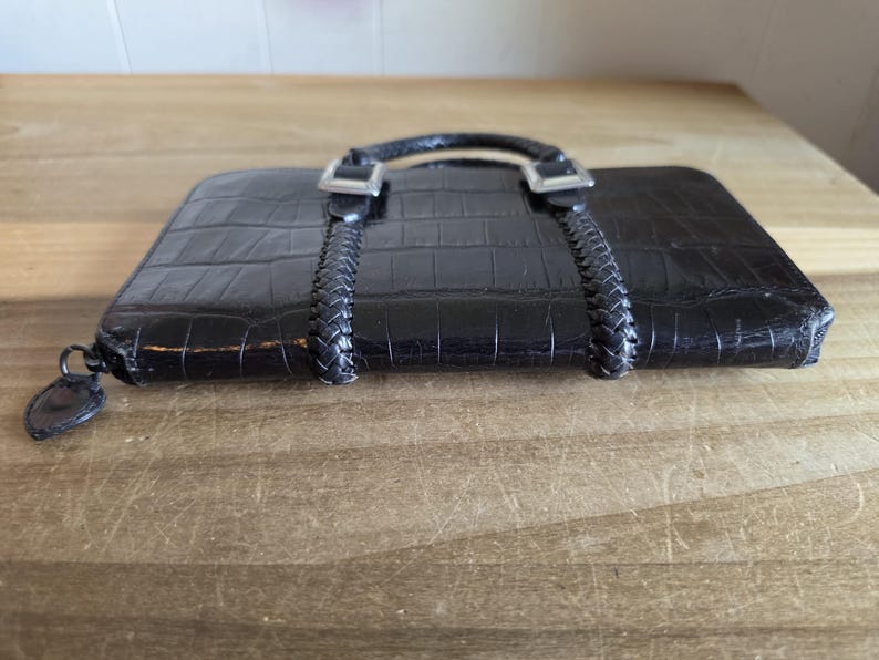 May include: A black rectangular wallet with a textured crocodile skin pattern. The wallet features two braided black straps extending from the top, secured by silver-toned metal buckles. A small, dark, heart-shaped zipper pull is attached to the side.