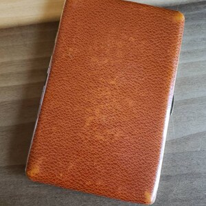 May include: A brown leather cigarette case with a silver metal frame. The case has a textured surface and a subtle embossed design.