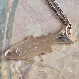 May include: A silver fish-shaped pendant necklace with a chain. The pendant has the text "Atlantic Lures Inc. Providence, R.I." engraved on it.