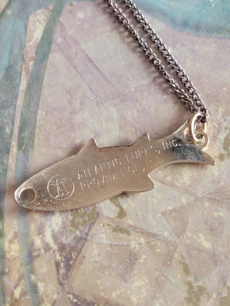 May include: A silver fish-shaped pendant necklace with a chain. The pendant has the text "Atlantic Lures Inc. Providence, R.I." engraved on it.