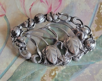 May include: Oval silver-toned brooch featuring a floral design with three prominent tulip-like blooms and surrounding buds. The brooch has a detailed, Art Nouveau style with curved lines and a polished finish. The background has a floral pattern.