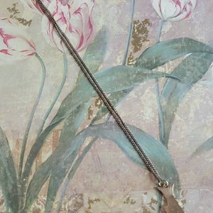 May include: A silver chain necklace with a silver fish pendant. The pendant is detailed with scales and fins. The necklace is photographed against a floral background.