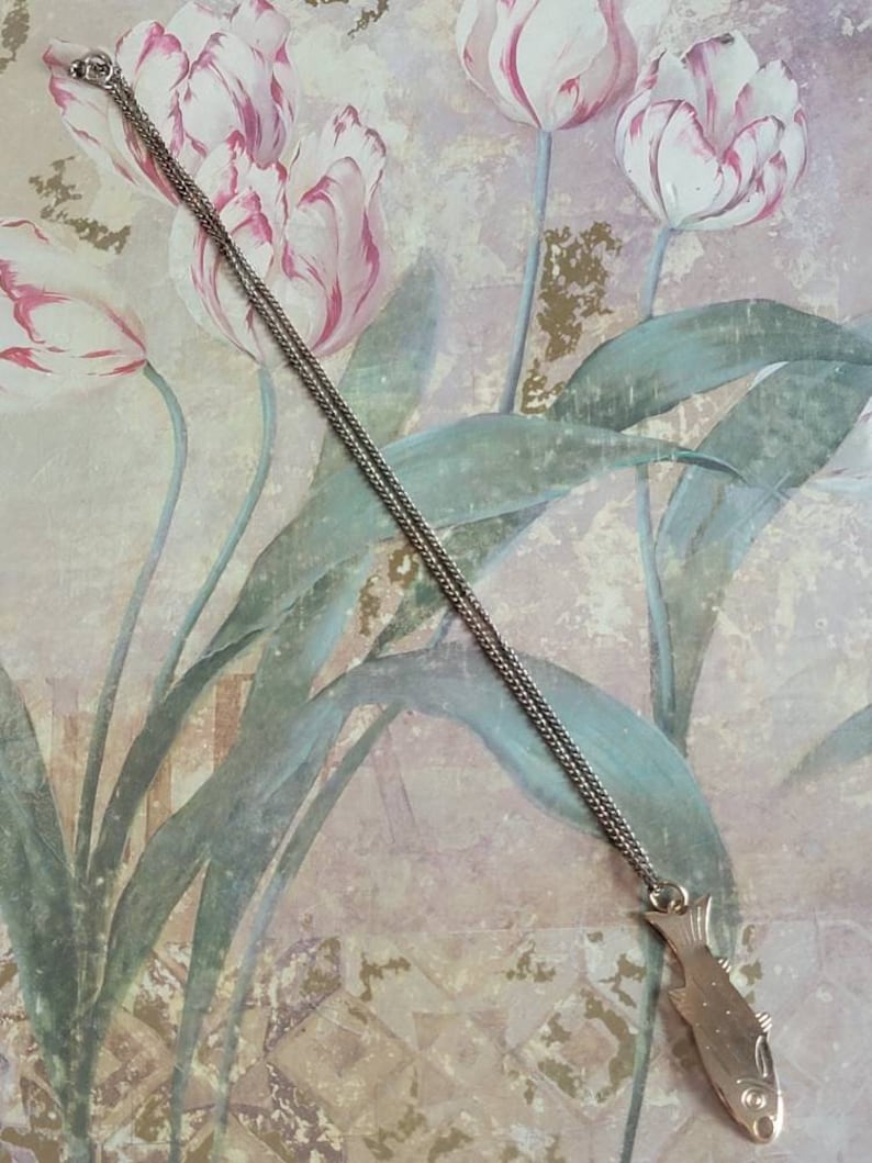 May include: A silver chain necklace with a silver fish pendant. The pendant is detailed with scales and fins. The necklace is photographed against a floral background.