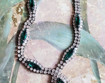 May include: A silver necklace with a teardrop-shaped pendant featuring a large emerald green stone surrounded by smaller clear rhinestones. The necklace is made of a chain of clear rhinestones with emerald green stones spaced throughout.