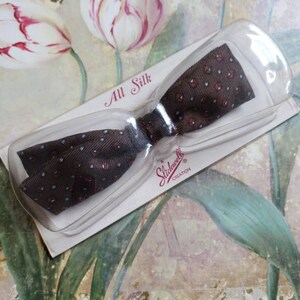 May include: A brown and white patterned bow tie made of silk. The bow tie is packaged in a clear plastic case with a white label that reads "All Silk" and "Slidewell Creation".