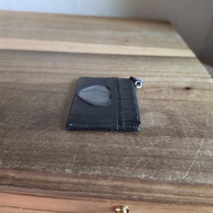 May include: A black leather card holder featuring a heart-shaped clear plastic window on the front. The accessory has a textured surface with a crocodile-embossed panel on the right side and a small metal zipper pull attached to the top corner.