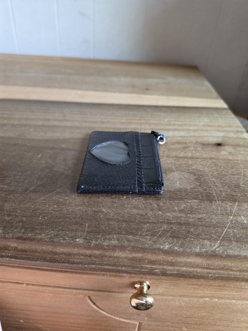 May include: A black leather card holder featuring a heart-shaped clear plastic window on the front. The accessory has a textured surface with a crocodile-embossed panel on the right side and a small metal zipper pull attached to the top corner.