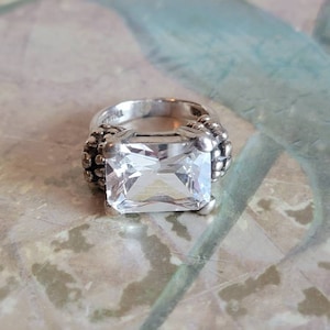 May include: A silver ring with a large, clear, square-shaped gemstone. The ring has a textured band with small beads.