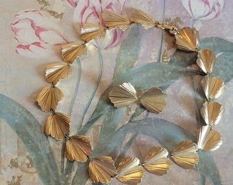 May include: A gold necklace with a fan-shaped design. The necklace is made of multiple pieces that are connected together. The necklace is on a floral background.