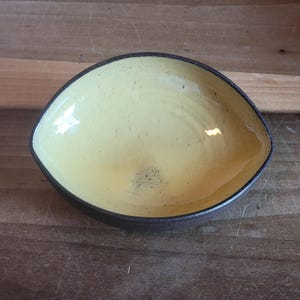 May include: A handmade, ceramic bowl with a unique, elongated shape. The interior is a pale yellow with subtle speckles, contrasting with the dark brown rim and exterior. This decorative bowl is ideal for serving snacks or as a small decorative piece.