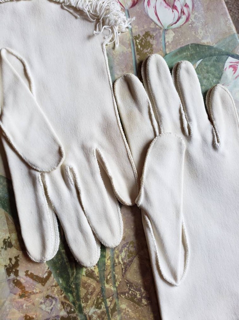 May include: A pair of white cotton gloves with a fringed trim. The gloves are long and have a delicate, feminine look.