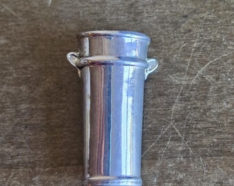 May include: A small, silver-toned metal miniature bucket or pail featuring a cylindrical body with a slightly flared rim and two small, decorative side handles. The surface has a smooth, reflective finish that catches the light.