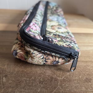 Puede incluir: Una bolsa rectangular con estampado floral y cierre de cremallera negro. La bolsa presenta un dise&ntilde;o tipo tapiz con flores rosas, azules, verdes y amarillas sobre un fondo beige. La cremallera es negra y la bolsa est&aacute; sobre una superficie de madera.