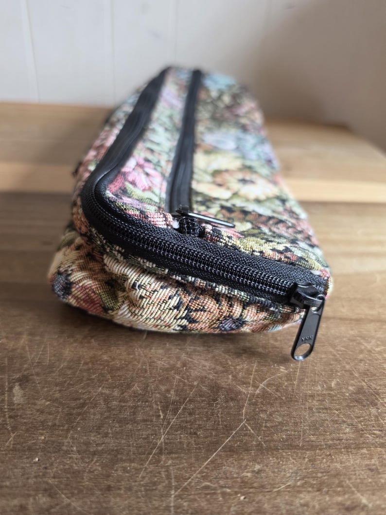 Puede incluir: Una bolsa rectangular con estampado floral y cierre de cremallera negro. La bolsa presenta un dise&ntilde;o tipo tapiz con flores rosas, azules, verdes y amarillas sobre un fondo beige. La cremallera es negra y la bolsa est&aacute; sobre una superficie de madera.