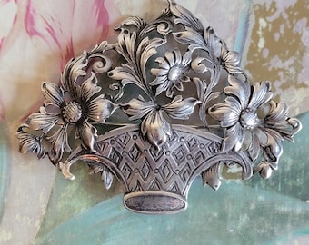 May include: A silver brooch depicting a basket brimming with flowers and leaves. The basket features a diamond design, and the flowers have detailed petals. The brooch is likely made of metal, with a vintage aesthetic.