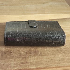 Puede incluir: Una cartera de cuero negra con un patr&oacute;n de cocodrilo en relieve y un cierre de solapa.