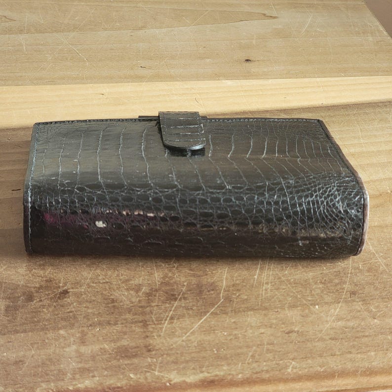 Puede incluir: Una cartera de cuero negra con un patr&oacute;n de cocodrilo en relieve y un cierre de solapa.