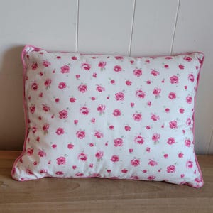 May include: Rectangular pillow with a white background covered in small pink rose patterns. The pillow has a pink border. The pillow is placed on a wooden surface.