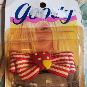 May include: A red and white striped bow with a red heart-shaped charm with yellow and green polka dots. The bow is packaged on a card with the word "Goody" printed in blue.