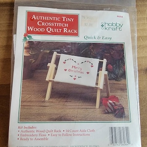 May include: A white cross stitch project with the words "Merry Christmas" in red on a wooden quilt rack. The kit includes 14 count Aida cloth, embroidery floss, and easy to follow instructions.