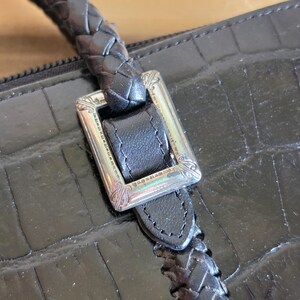 May include: A black handbag with a crocodile-embossed texture, featuring a braided leather handle attached to the body with a rectangular silver-toned metal buckle that has engraved decorative details along its frame.