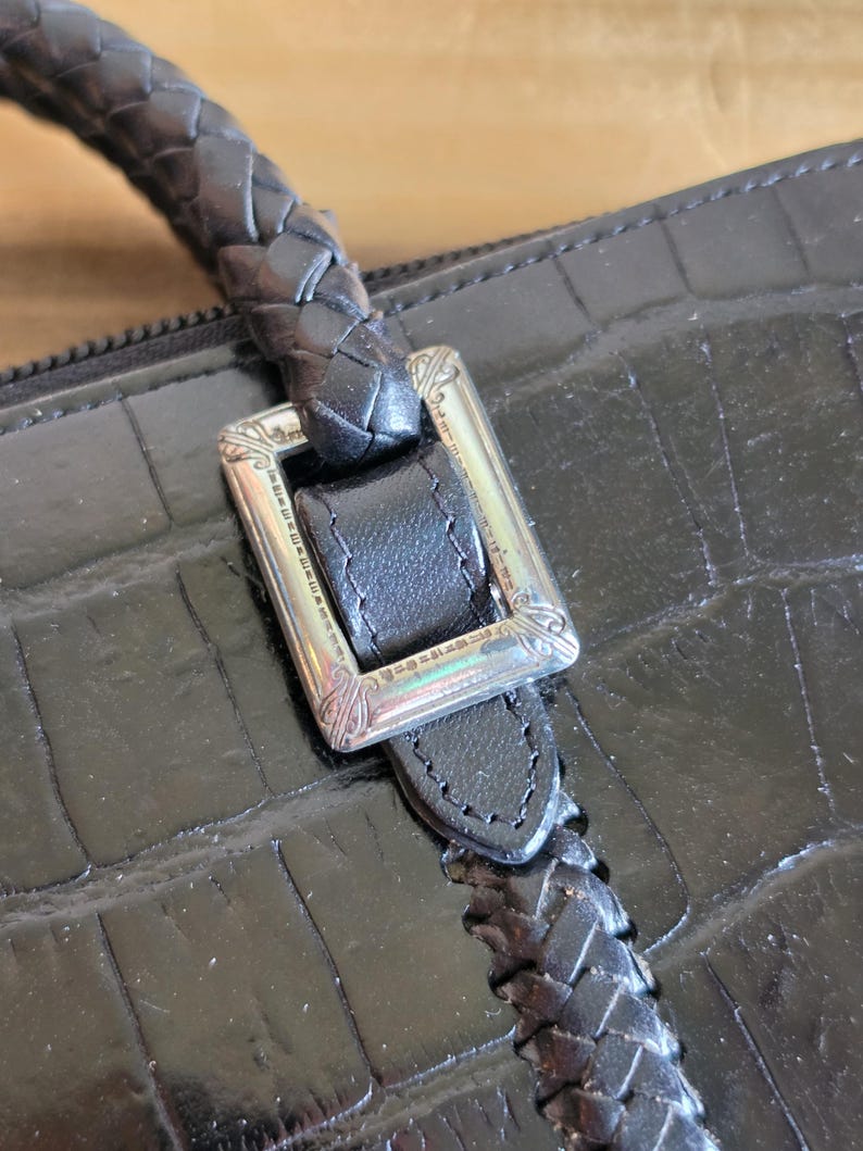 May include: A black handbag with a crocodile-embossed texture, featuring a braided leather handle attached to the body with a rectangular silver-toned metal buckle that has engraved decorative details along its frame.