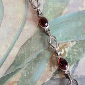 May include: Silver-toned chain bracelet with oval, reddish-brown gemstones. The bracelet features a series of knot-like links connecting the gemstones. The background has a vintage, painted appearance with muted colors.