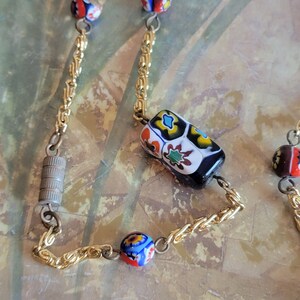 May include: A gold chain necklace with colorful glass beads. The beads are round and rectangular, with intricate patterns in shades of red, blue, green, yellow, and white.