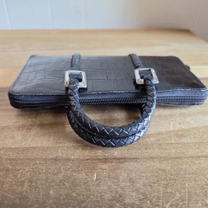 May include: A black rectangular wallet with a crocodile-embossed texture, featuring a zipper closure and two braided handles attached with silver-toned metal hardware. The wallet has a structured design with a smooth finish on the top surface.