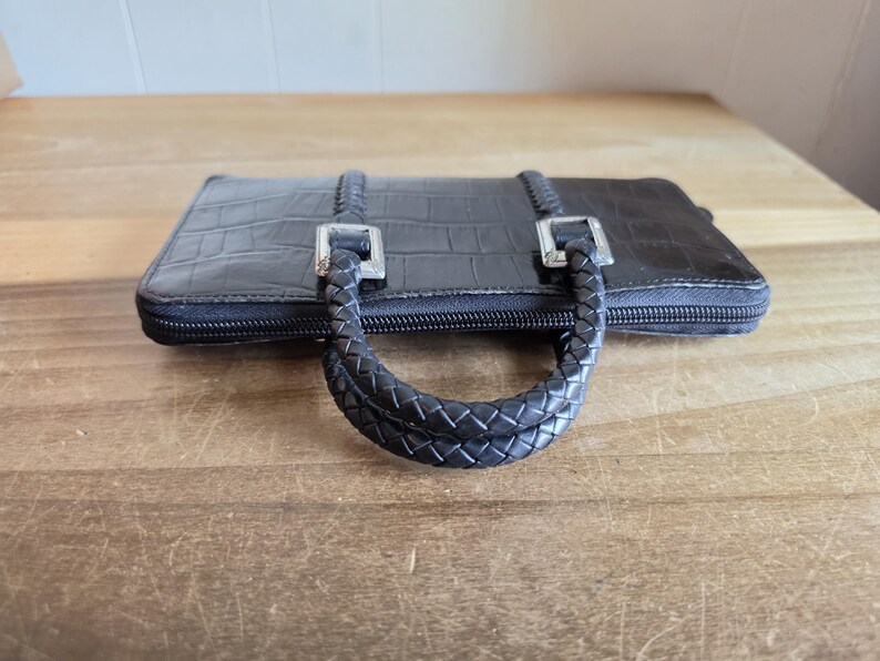 May include: A black rectangular wallet with a crocodile-embossed texture, featuring a zipper closure and two braided handles attached with silver-toned metal hardware. The wallet has a structured design with a smooth finish on the top surface.