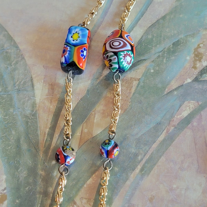 May include: A gold chain necklace with colorful glass beads. The beads are round and rectangular, with intricate patterns of flowers and swirls. The necklace is a statement piece with a vintage aesthetic.