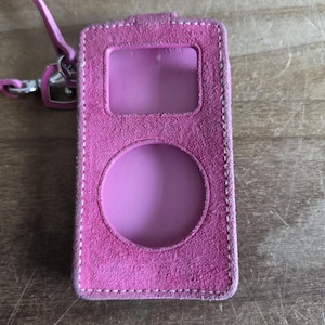 May include: A bright pink suede protective case for a portable media player, featuring a rectangular screen cutout and a circular click wheel cutout. The case includes white contrast stitching along the edges and a matching pink wrist strap with a silver metal heart-shaped clasp attached to the top.