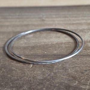 May include: Two thin, circular metal bangles with a dark, oxidized finish. The bracelets feature a subtle hammered texture along the surface, creating a slightly uneven, reflective appearance. The two rings are interlocked, forming a single unit.