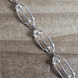 May include: A silver-toned chain necklace with a repeating open oval and ball design. The necklace is displayed on a wooden surface, showcasing its intricate links and overall aesthetic.