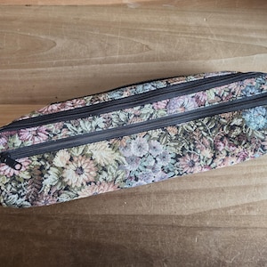 Puede incluir: Un estuche rectangular de tapiz floral con tres cremalleras negras. El estuche presenta un estampado de flores en tonos rosas, verdes y beige. El estuche est&aacute; sobre una superficie de madera.
