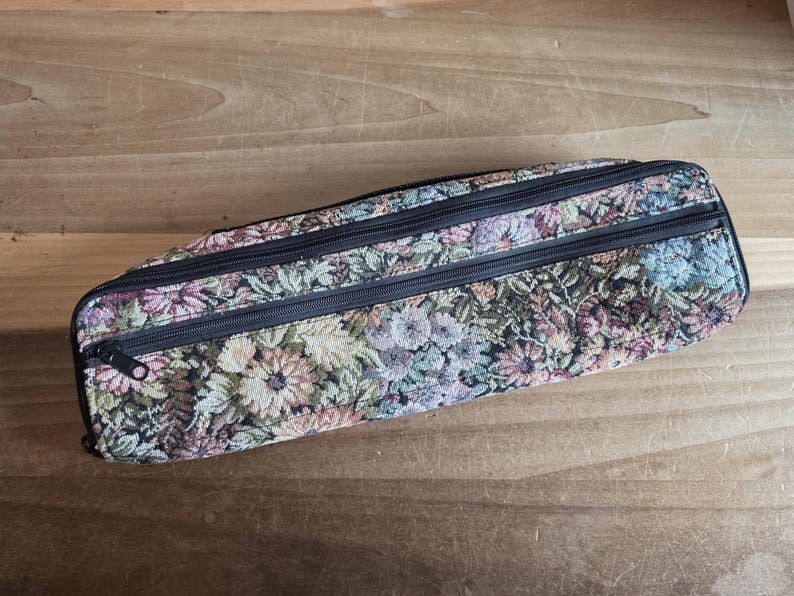 Puede incluir: Un estuche rectangular de tapiz floral con tres cremalleras negras. El estuche presenta un estampado de flores en tonos rosas, verdes y beige. El estuche est&aacute; sobre una superficie de madera.