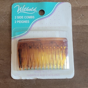 May include: A package containing two brown plastic combs. The package is white with blue and pink accents. The text on the package reads "Wilhold", "2 Side Combs", "2 Peignes", "Made in U.S.A.", "Fabrique aux Etats-Unis", and "1751".
