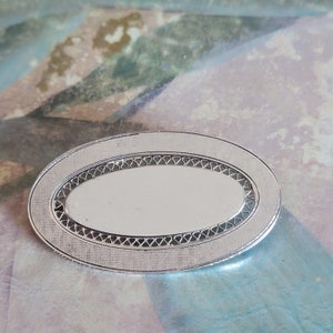 May include: A silver oval-shaped brooch with a decorative border and a smooth, reflective center.