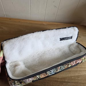 Puede incluir: Estuche rectangular abierto con exterior de tapiz floral y forro interior blanco y suave. El estuche tiene una cremallera negra y una etiqueta que dice "Pearl Flute F 100". El estuche est&aacute; abierto, revelando el interior de felpa.