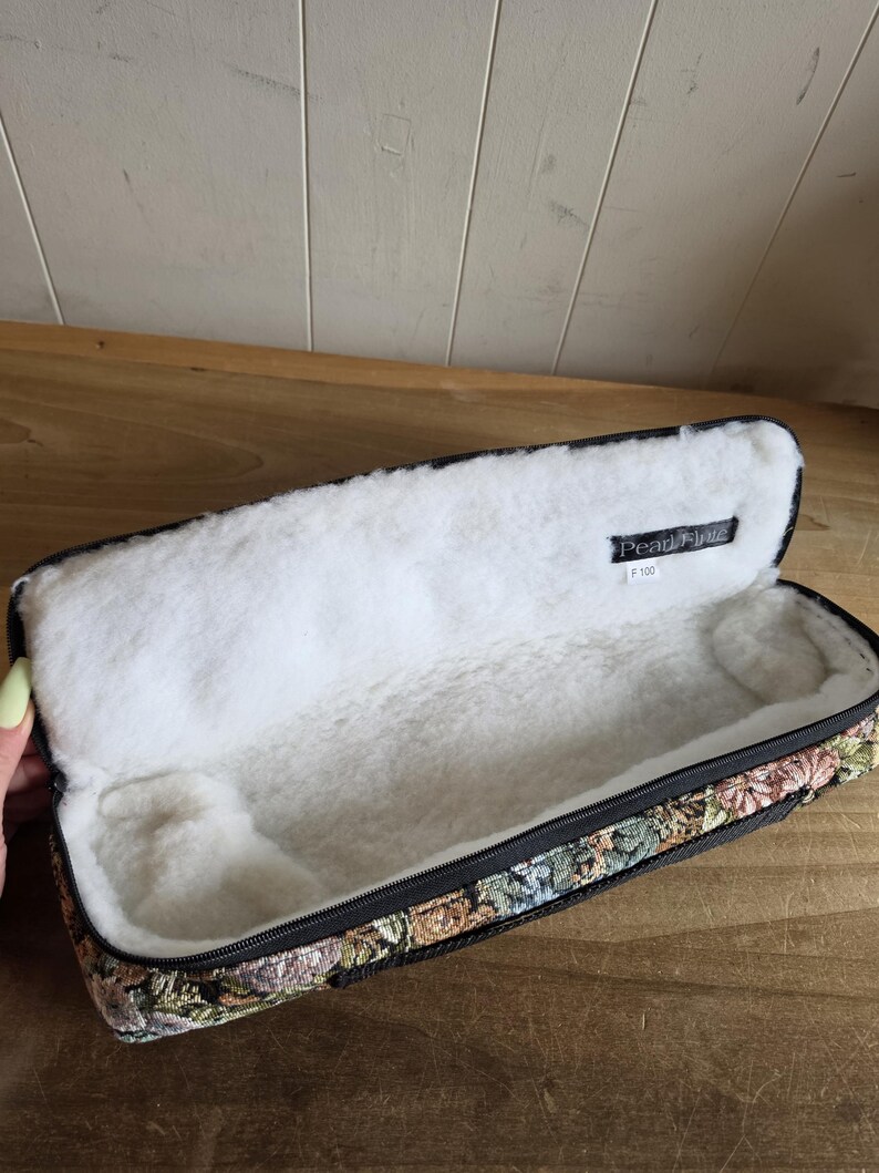 Puede incluir: Estuche rectangular abierto con exterior de tapiz floral y forro interior blanco y suave. El estuche tiene una cremallera negra y una etiqueta que dice "Pearl Flute F 100". El estuche est&aacute; abierto, revelando el interior de felpa.