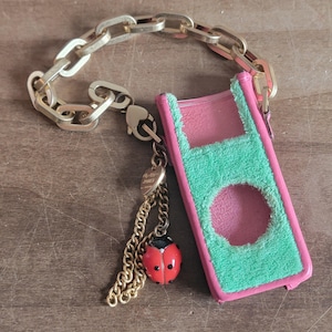 May include: A pink and green iPod case with a gold chain and a ladybug charm. The case has a pink interior and a green, textured exterior. The ladybug charm is red with black spots.
