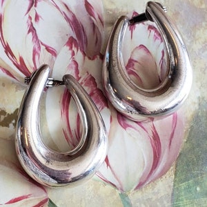 May include: A pair of silver-toned hoop earrings. The earrings have a teardrop shape and a polished finish. The background features a floral pattern with pink and white tulips.