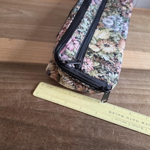 Puede incluir: Un estuche rectangular con estampado floral y cremalleras negras. El estuche est&aacute; hecho de una tela con un estampado de flores rosas, azules y verdes. Una regla amarilla est&aacute; al lado del estuche.