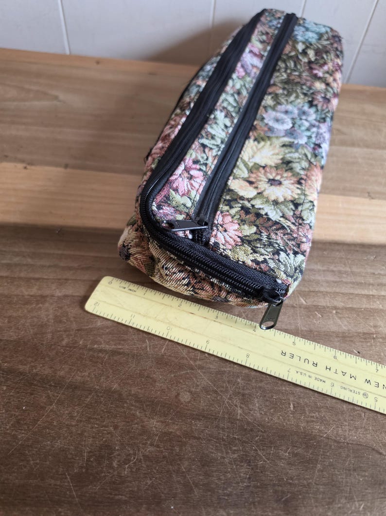 Puede incluir: Un estuche rectangular con estampado floral y cremalleras negras. El estuche est&aacute; hecho de una tela con un estampado de flores rosas, azules y verdes. Una regla amarilla est&aacute; al lado del estuche.