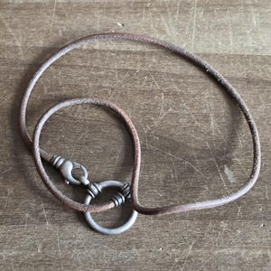 May include: A brown leather necklace with a silver-colored clasp and a large silver-colored ring. The necklace is lying on a wooden surface.