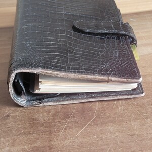 Puede incluir: Un diario encuadernado en cuero negro con un patr&oacute;n texturizado de piel de cocodrilo. El diario tiene un cierre de solapa con un broche met&aacute;lico.
